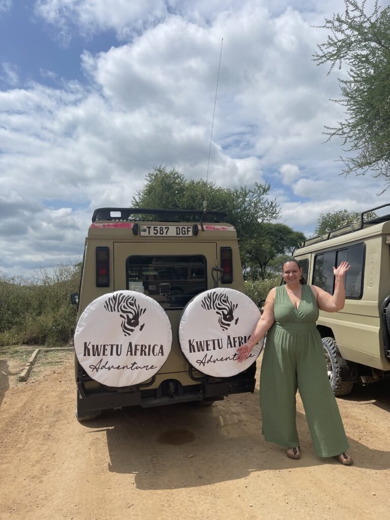 A happy tourist posing next to a KwetuAfrica Adventure safari vehicle, showcasing the company's logo on the spare tire covers. The scene is set on a bright, sunny day with a blue sky and fluffy clouds in the background.