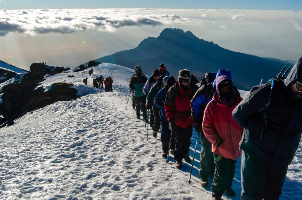 A group of climbers making their way up the snowy slopes of Mount Kilimanjaro, with stunning mountain peaks and a sea of clouds in the background. The climbers are dressed warmly and using trekking poles, highlighting the challenging yet rewarding nature of the ascent.