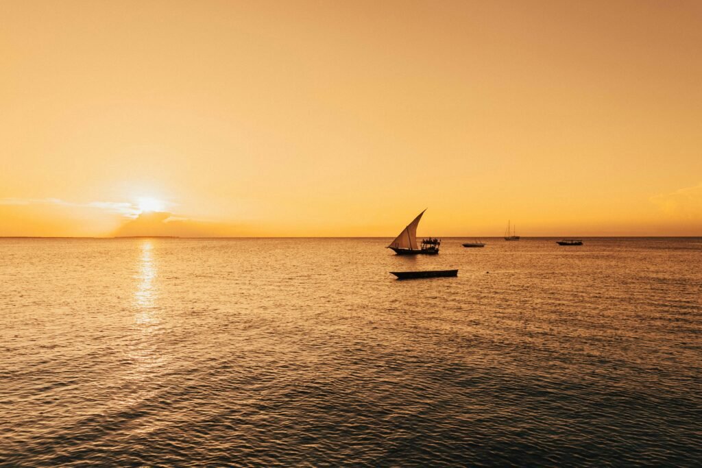 A traditional dhow sailing boat glides across the calm waters of Zanzibar at sunset. The sky is painted in golden hues, reflecting beautifully on the ocean surface, creating a serene and picturesque scene.