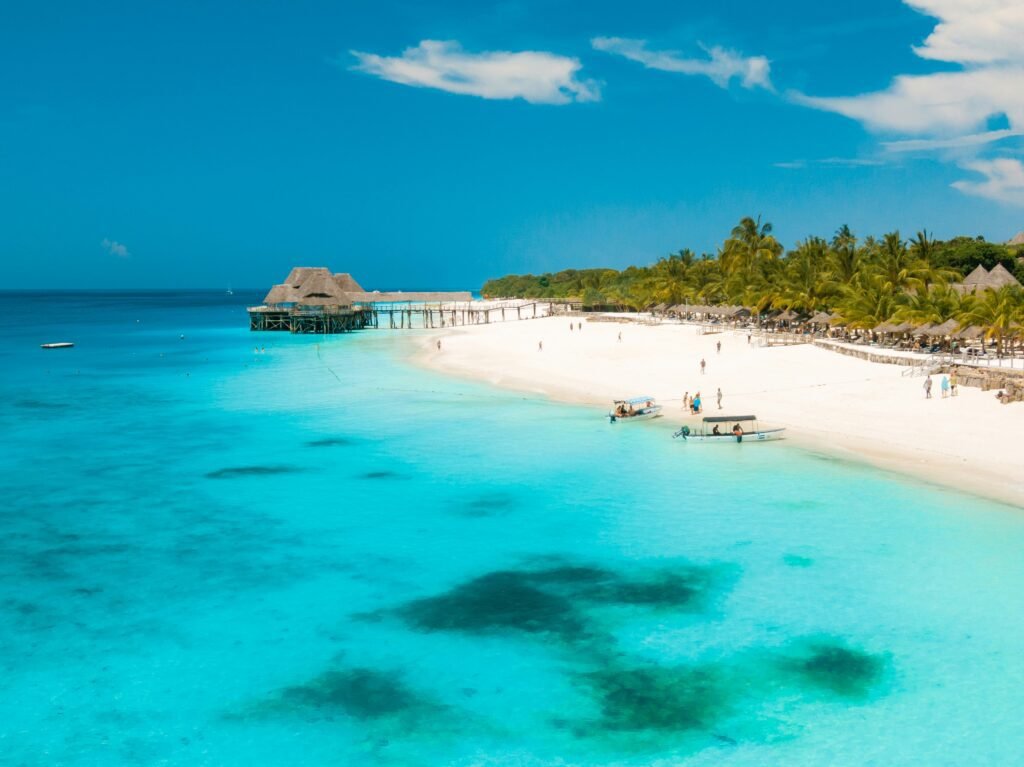 A stunning beach in Zanzibar with turquoise waters, white sand, and a pier extending into the ocean. Palm trees line the shore, providing shade to visitors relaxing on sun loungers. Small boats are anchored near the shore, highlighting the serene and inviting atmosphere of this tropical paradise.