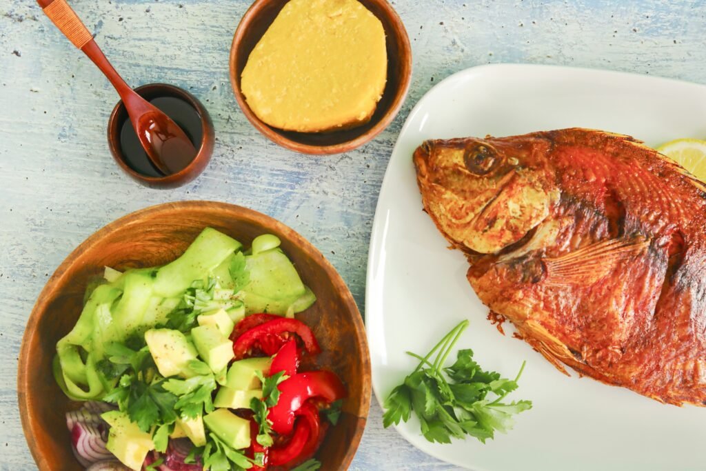 A traditional Tanzanian meal featuring a beautifully grilled fish served on a white plate, accompanied by a fresh salad with avocado, tomatoes, and cucumber in a wooden bowl. Nearby are a bowl of creamy polenta and a small dish of sauce, presented on a rustic blue wooden table.