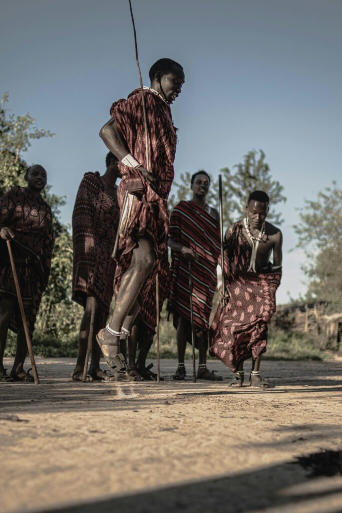 Maasai warriors engaging in a traditional jumping dance, dressed in their vibrant red and patterned shukas. The scene captures the cultural heritage and energetic spirit of the Maasai tribe against a backdrop of trees and a clear sky.