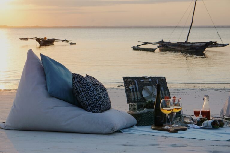 A cozy beach picnic setup with cushions, a picnic basket, and wine glasses at sunset in Zanzibar. Traditional wooden boats float on the calm waters in the background, creating a serene and romantic atmosphere.