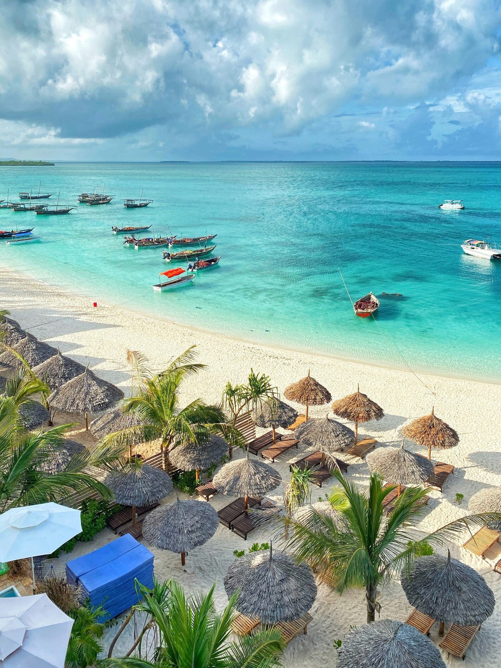 A picturesque view of a tropical beach in Zanzibar, featuring turquoise waters, traditional wooden boats, and rows of thatched umbrellas on the white sandy shore. Palm trees and lounge chairs add to the idyllic and serene atmosphere of this coastal paradise.