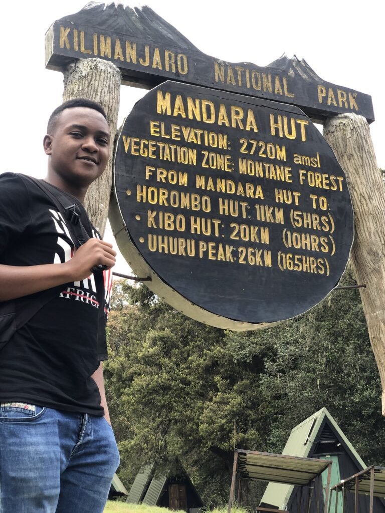 A hiker standing next to the Mandara Hut sign in Kilimanjaro National Park. The sign indicates the elevation of 2720 meters above sea level and provides distances and estimated hiking times to Horombo Hut, Kibo Hut, and Uhuru Peak. The background shows the lush montane forest and hut structures.