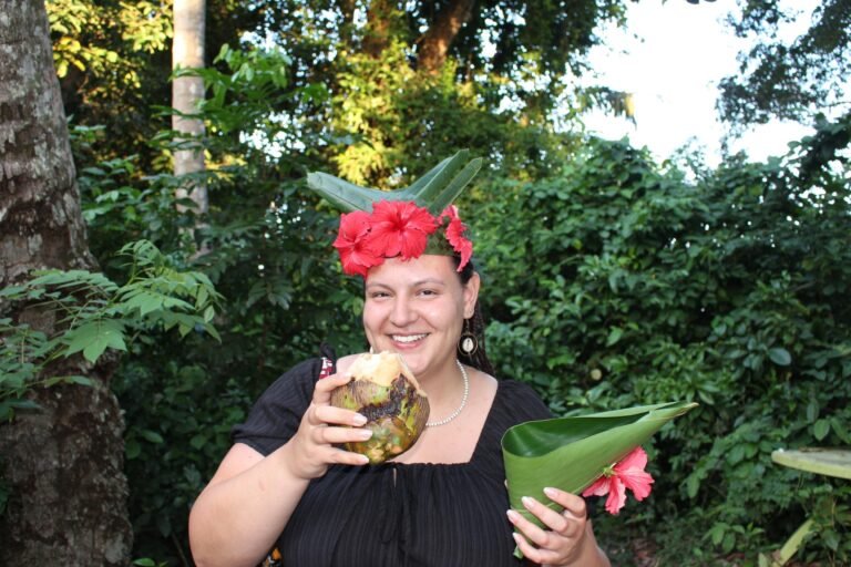 A joyful woman, adorned with a handcrafted crown of leaves and red hibiscus flowers, smiles brightly while holding a fresh coconut and a folded leaf in her hands. She stands amidst lush, green foliage, embodying the spirit of cultural immersion and the natural beauty of Tanzania.