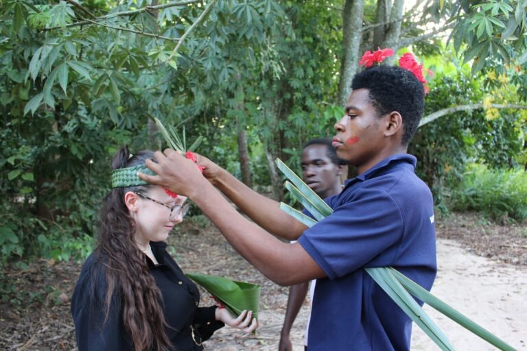 A local guide decorates a tourist with a handmade plant crown in a lush forest setting, offering a glimpse of Tanzanian cultural traditions, as another local observes.