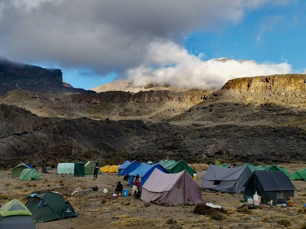 Photo of a mountain base camp with colorful tents set up on rocky terrain, surrounded by dramatic cliffs and partially shrouded in clouds under a bright blue sky.