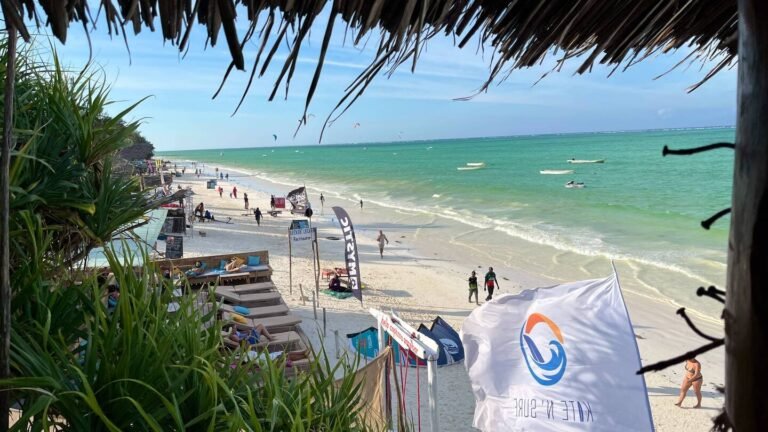 A view from a thatched roof, overlooking the vibrant beach in Zanzibar, with kite surfers, beachgoers, and local shops along the sandy shore, framed by lush greenery.