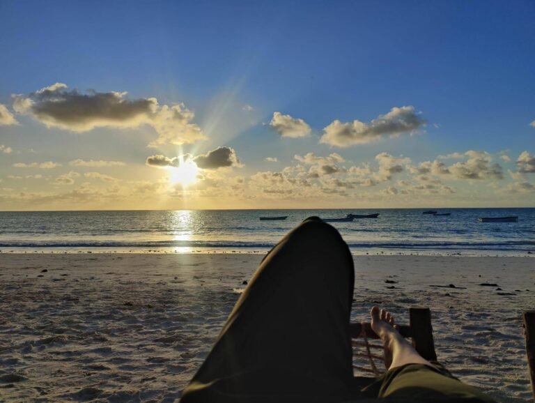 A peaceful view of the beach at sunset, with boats floating on the calm water and a person reclining in the foreground, enjoying the serene ambiance of the moment.