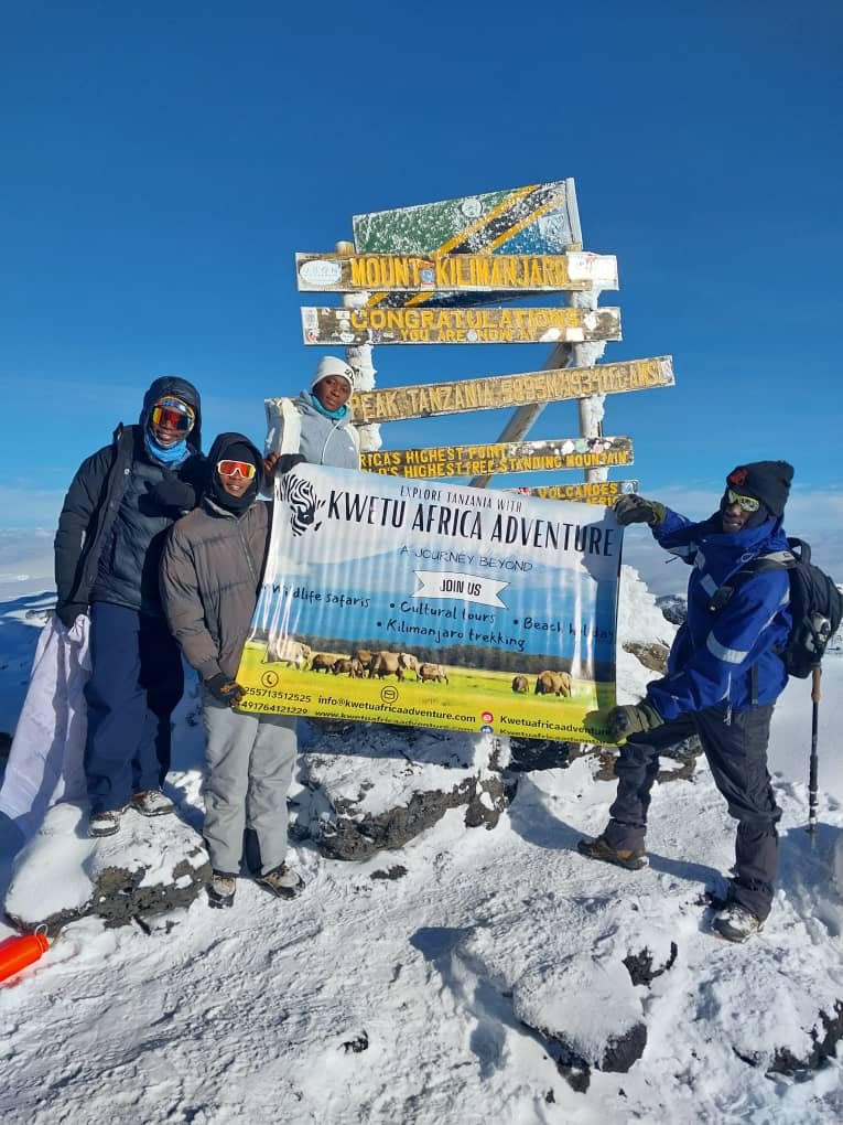 A group of climbers celebrates at the summit of Mount Kilimanjaro with a banner for KwetuAfrica Adventure, showcasing the breathtaking snowy landscape and a sign marking Africa’s highest peak.