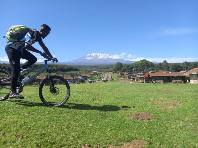 A cyclist rides through a rural village with the majestic snow-capped peak of Mount Kilimanjaro in the distance, under a bright blue sky.