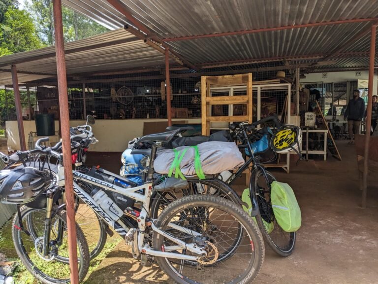 Two bikes are parked outside a rustic building, ready for the next leg of the journey, with travel gear strapped to them and a glimpse of the local environment in the background.