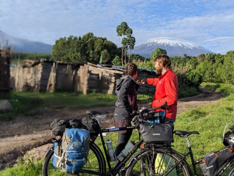 Two cyclists take a moment to chat while their bikes are parked, with Mount Kilimanjaro towering in the background, offering a stunning view of the snow-capped peak.