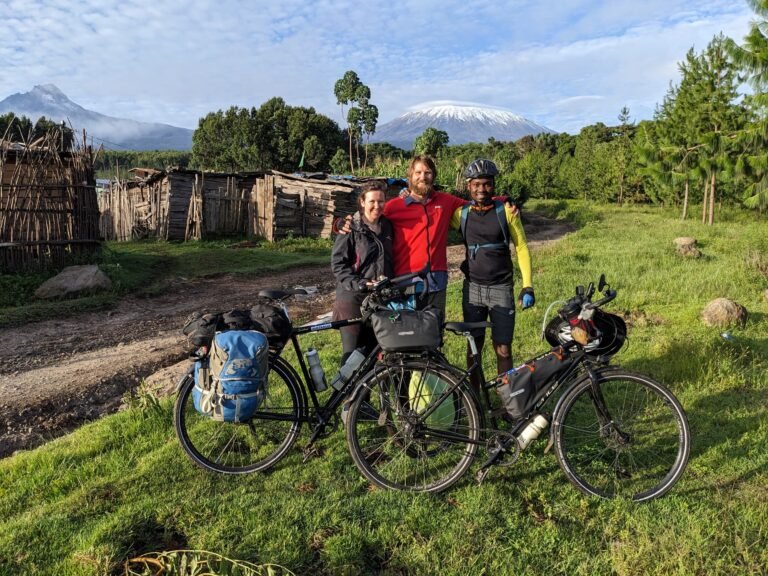 Three cyclists with bikes loaded with gear stand together in front of Mount Kilimanjaro, surrounded by a rural Tanzanian landscape.