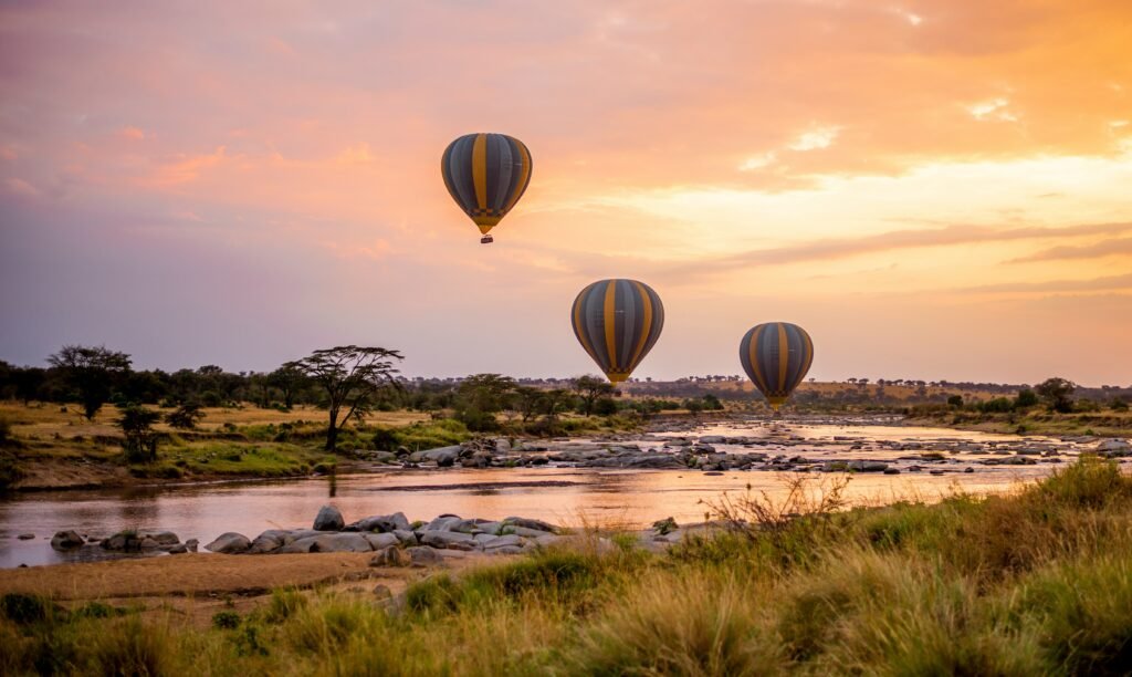 Photo of hot air balloons floating gracefully over a river at sunrise, with golden skies and a scenic savannah landscape below.